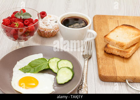 Plate with fried egg, fresh cut cucumber and leaf of basil, glass bowl with fresh raspberries, cup of black coffee, fork, muffin and toasts on wooden  Stock Photo