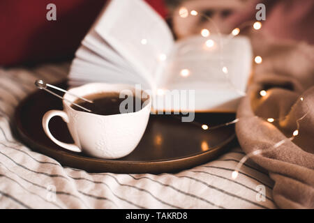 Cup of balck tea with wooden tray over lights and open book in bed closeup. Good morning. Stock Photo