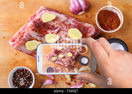 hands of man with smartphone taking photo Raw fresh meat, uncooked lamb or beef ribs with pepper, garlic, salt and spices on dark stone background Stock Photo