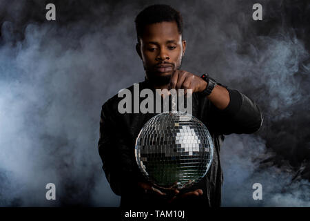 Confident brutal dark skinned man holding disco ball, looking at camera, going to party with friends, wearing casual clothes standing over black backg Stock Photo
