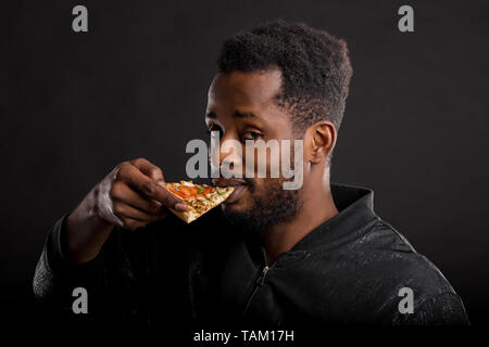 Closeup portrait of hungry unshaven african american guy with piece of tasty pizza in his hand, eating fast food isolated on black background. Unhealt Stock Photo