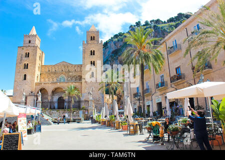 Cefalu, Sicily, Italy - Apr 7th 2019: Tourists on the old town square in the outdoor cafe gardens in front of Cefalu Cathedral. Famous Roman Catholic Basilica in Norman style is popular attraction. Stock Photo