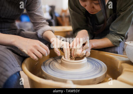 Close-up of skilled Asian female potter showing woman how to mould clay dish using pottery wheel in workshop Stock Photo