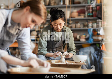 Serious skilled young Asian female potter in apron sitting at pottery wheel and making clay vase with hands in workshop Stock Photo