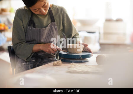 Serious concentrated young Asian woman in apron sitting at table and using cutter while making bowl in workshop Stock Photo