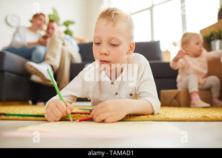 Serious blond-haired boy in casual sweater lying on soft carpet and using colorful pencil while drawing picture at home Stock Photo