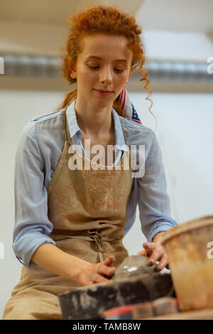 Carving new pot. Pleasant good-looking ginger lady in light shirt sitting at the pottery wheel and creating new form Stock Photo