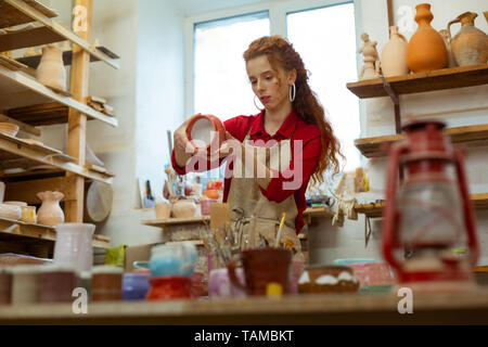 Focused ginger lady. Curious good-looking curly woman in red shirt observing finished pot while organizing studio Stock Photo