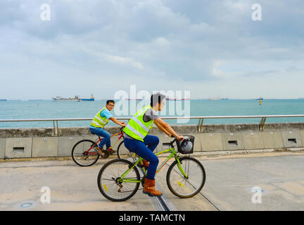 SINGAPORE - FEBRUARY 16, 2107: Two worker in uniform riding the bicycles at Singapore harbor. Industrial cargo ships in the background Stock Photo