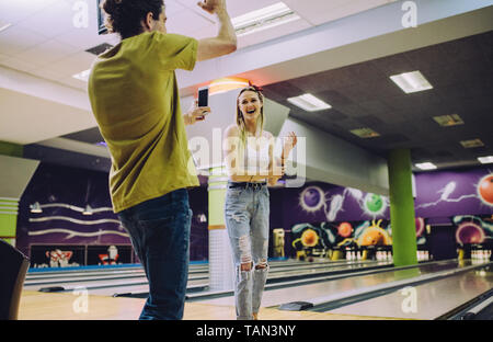 Young man recording woman playing bowling at club. Young friends enjoying playing at bowling arena. Stock Photo