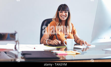 Illustrator making a sketch on a digital writing pad. Smiling woman entrepreneur working on her designs sitting at her desk in office. Stock Photo