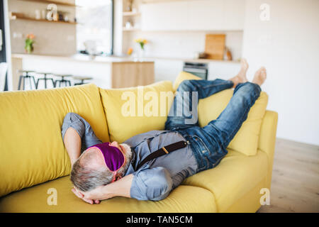 Senior man with eye mask lying on sofa indoors at home, relaxing. Stock Photo