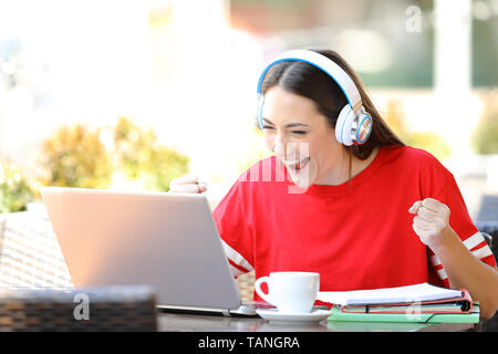 Excited student e-learning with a laptop and headphones finds offers in a coffee shop terrace Stock Photo