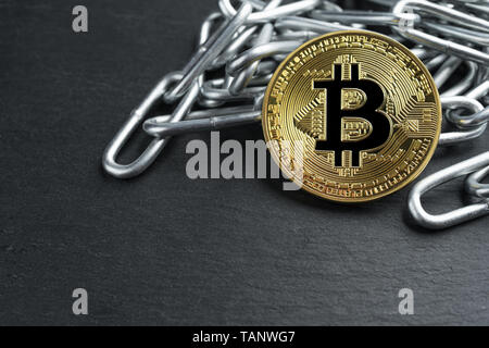 Physical Bitcoin gold coin on a convoluted shiny metal chain on dark stone slate plate background. Cryptocurrency legal restrictions abstract concept. Stock Photo