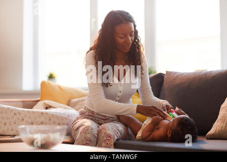 Smiling attractive curly-haired young black mother in homewear sitting on sofa in living room and dressing baby boy while taking care of him Stock Photo