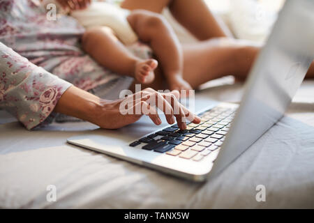 Close-up of unrecognizable African mother sitting on bed and browsing internet on laptop while baby sleeping in her arm Stock Photo