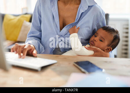 Close-up of concentrated African baby boy keeping mothers shirt and looking at computer monitor while his mother working on project Stock Photo