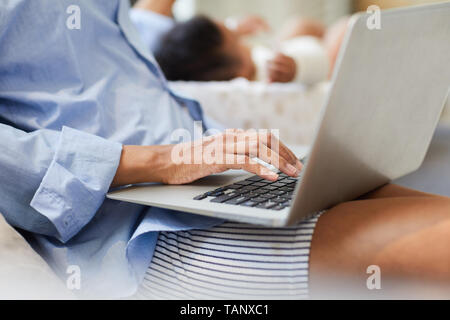 Close-up of unrecognizable clack mother in oversize shirt sitting on sofa and checking files on laptop while baby sleeping in background Stock Photo