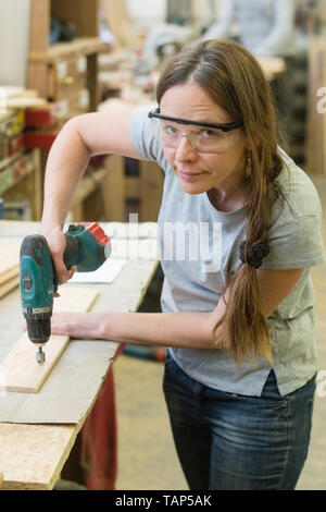 Young woman with drill making hole into wooden plank at workshop Stock Photo
