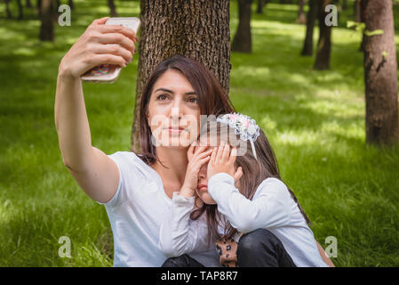 Young mom and little girl takes selfie together.Little girl hides her face.Photo of young mother and her daughter having selfie on green grass Stock Photo