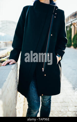 Unrecognizable young young woman wearing dark blue jeans and black coat standing outdoors leaning on parapet on sunny spring day. Stock Photo