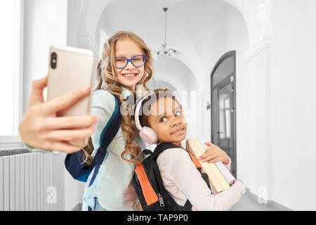 Beautiful, funny school friends doing selfie in school hallway on phone. Adorable girl with long hair in glasses holding phone. Little child holding books. Two schoolgirls looking at gadget.