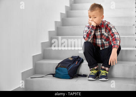 Handsome, thoughtful schoolboy sitting on stairwell in school, leaning head on hand. Lonely, sad, depressed child in checked shirt, jeans and sneakers thinking. Stock Photo