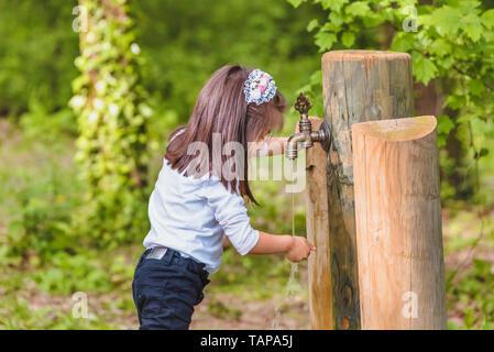 Adorable four years old cute little girl drinks water from a wooden tap in forest at a sunny day Stock Photo