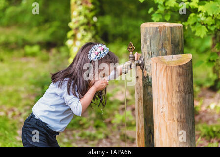 Adorable four years old cute little girl drinks water from a wooden tap in forest at a sunny day Stock Photo