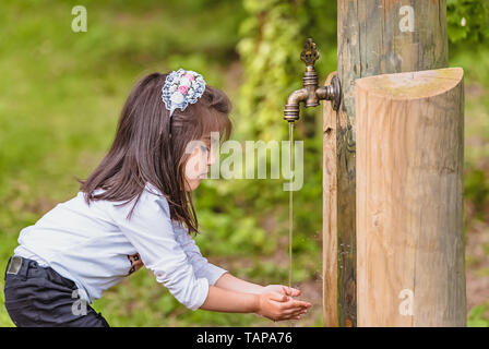 Adorable four years old cute little girl drinks water from a wooden tap in forest at a sunny day Stock Photo