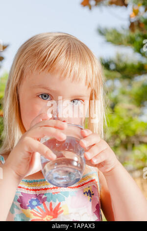 Blonde little girl drinking fresh and pure tap water from a glass in a summer garden - healthy lifestyle concept Stock Photo