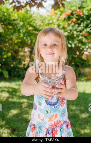 Blonde little girl holding glass of fresh and pure tap water in a summer garden, focus on glass of water Stock Photo