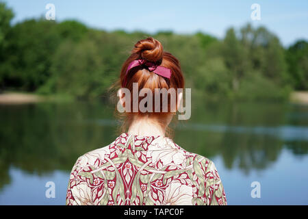 back view of unrecognizable young woman with floral dress and red hair bun looking at lake in solitude Stock Photo