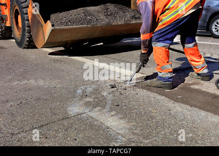 A road maintenance worker removes old asphalt on the roadway with a jackhammer into an excavator during road construction. Stock Photo