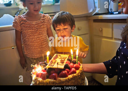 Young boy blowing out his birthday candles as his smiling mother holds the birthday cake in their kitchen. Stock Photo