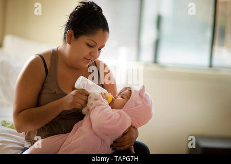 Young mother sitting on the bed giving her baby daughter a bottle. Stock Photo