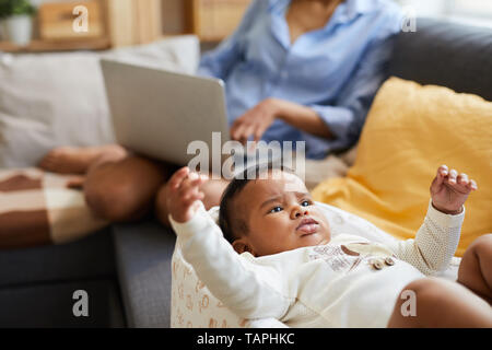 Close-up of angry frowning cute black baby boy in bodysuit lying in cocoon and gesturing hands while mother working in background Stock Photo