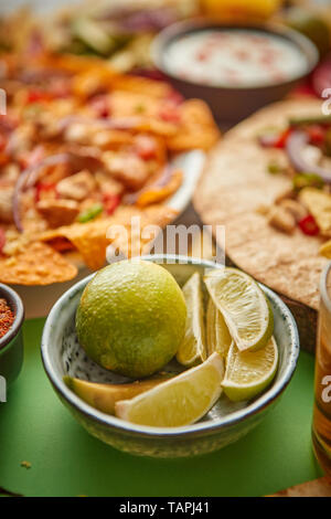 Close up on lime slices in ceramic bowl with various freshly made Mexican foods assortment in the back. Placed on colorful table. With nachos, tacos,  Stock Photo