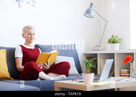 Blonde middle aged woman in casualwear sitting on couch and reading interesting book of stories at leisure Stock Photo