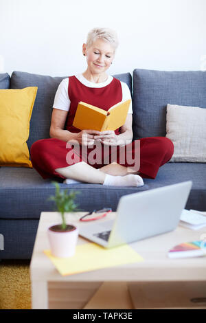 Happy middle aged female with book in hands sitting on comfortable couch in front of small table and laptop and reading at home Stock Photo