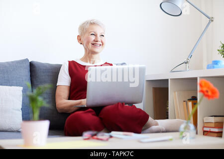 Mature blonde cheerful woman with laptop relaxing on couch at home at leisure and laughing at something Stock Photo