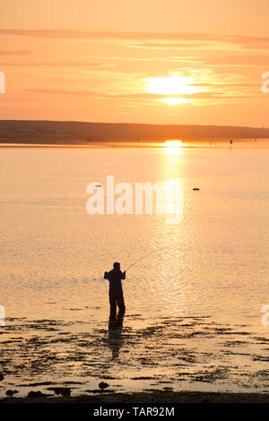 A man saltwater flyfishing in the Fleet lagoon at sunset viewed from the bridge at Ferry Bridge. Ferry Bridge is at the start of the causeway that lea Stock Photo