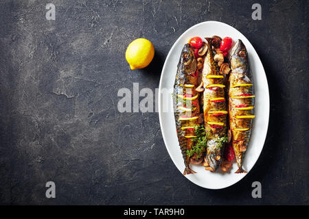 three whole broiled mackerels with lemon, tomatoes, mushrooms, spices and herbs on a white oval dish, horizontal view from above, flatlay, copy space Stock Photo