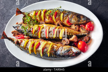 close-up of three whole broiled mackerels with lemon, tomatoes, mushrooms, spices and herbs on a white oval dish, horizontal view from above Stock Photo