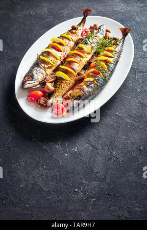 three whole broiled mackerels with lemon, tomatoes, mushrooms, spices and herbs on a white oval dish, vertical view from above, copy space Stock Photo