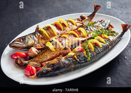 close-up of three whole broiled mackerels served with lemon, tomatoes, mushrooms, spices and herbs on a white oval plate, horizontal view from above Stock Photo