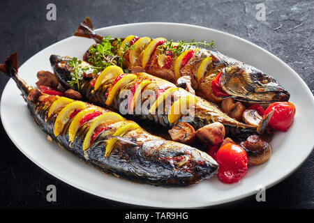 close-up of three whole broiled mackerels served with lemon, tomatoes, mushrooms, spices and herbs on a white oval platter, horizontal view from above Stock Photo