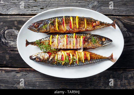 close-up of three whole grilled mackerel with lemon slices and fresh thyme on a white oval platter on an old wooden table, horizontal view from above, Stock Photo