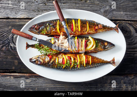 close-up of three whole grilled mackerel with lemon slices and fresh thyme on a white oval platter with fork and knife on an old wooden table, horizon Stock Photo