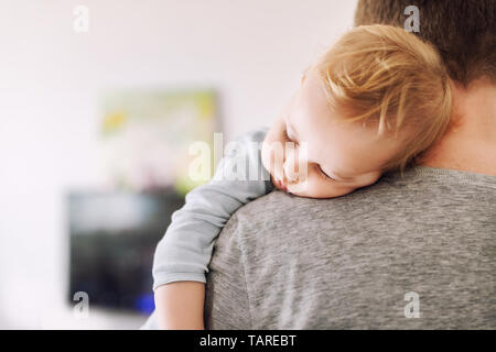 Close-up portrait of cute adorable blond caucasian toddler boy sleeping on fathers shoulder indoors. Sweet little child feeling safety daddys hand Stock Photo